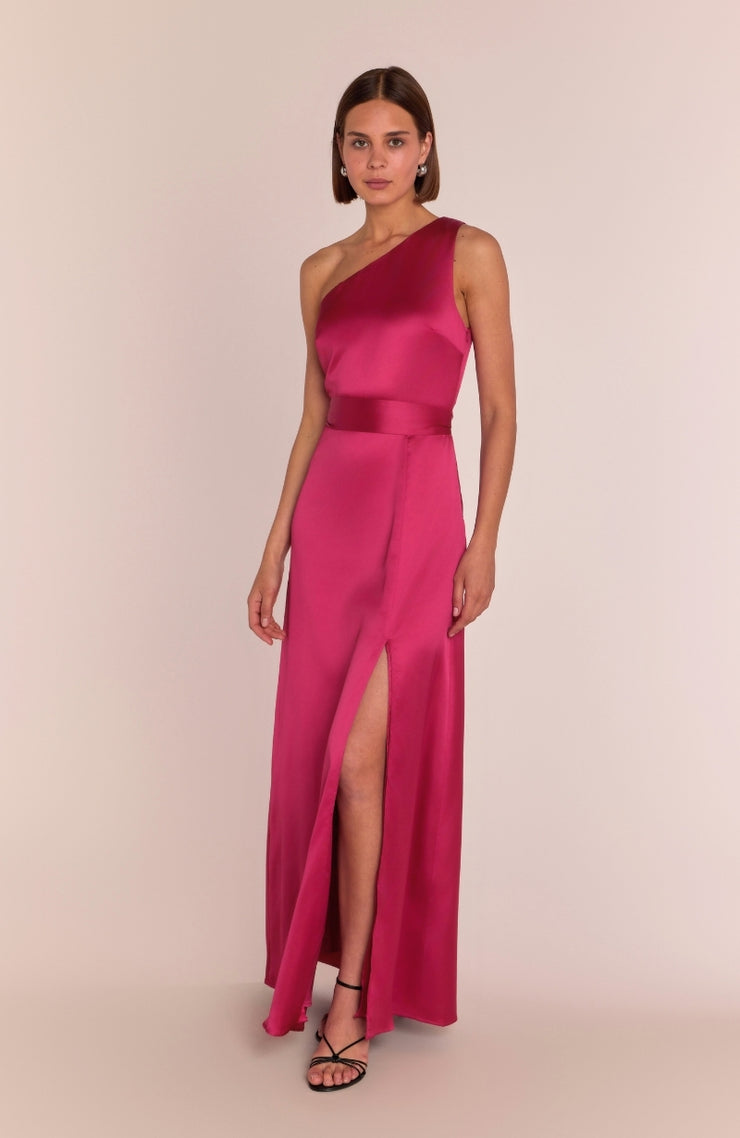 A model wearing a hot pink satin one-shoulder floor-length dress with a sash and a slit, standing against a pink background.