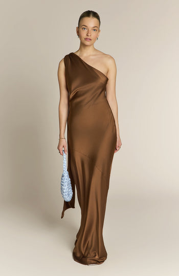 Woman wearing a brown one-shoulder dress holding a blue clutch against a beige background